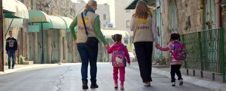 Zwei Ökumenische Begleiter:innen begleiten zwei kleine Mädchen auf dem Weg zur Schule.