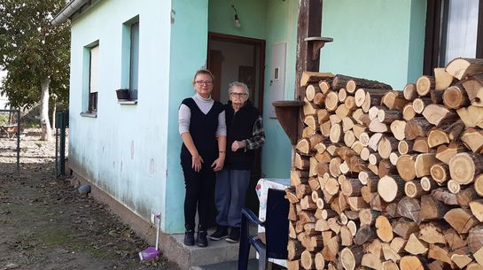 Eine ältere und eine jüngere Frau in einem Hauseingang, neben einem großen Holzstoss.