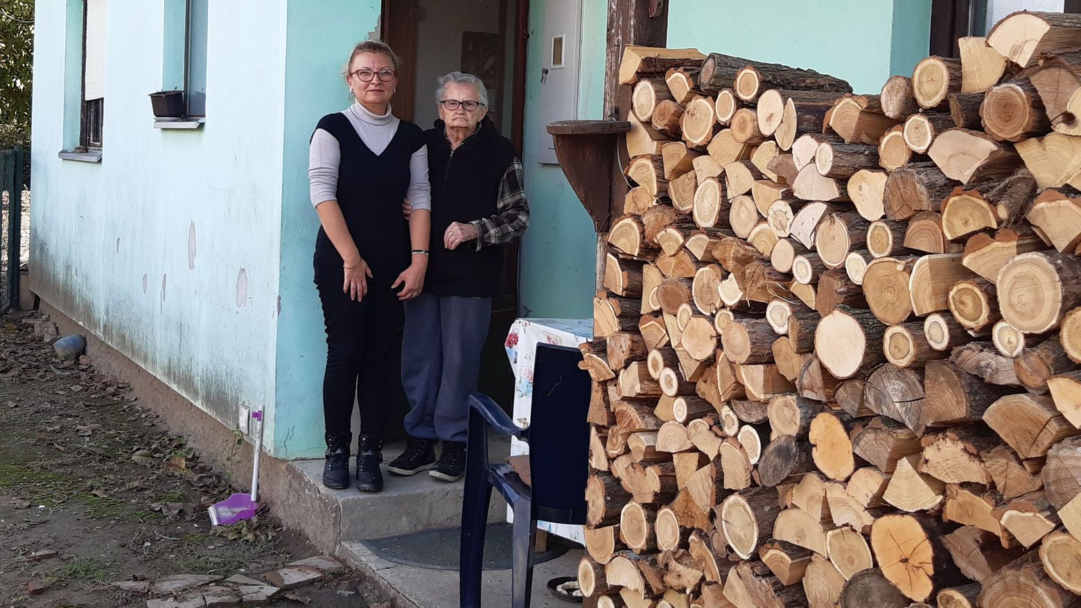 Eine ältere und eine jüngere Frau in einem Hauseingang, neben einem großen Holzstoss.