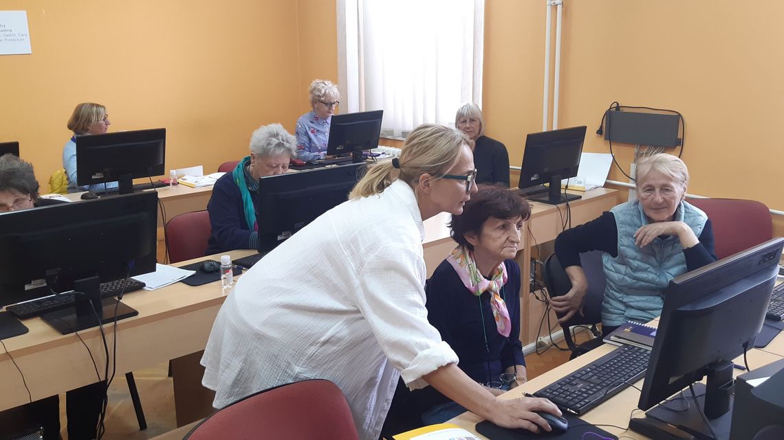Ältere Menschen sitzen an Tischen vor Computern, eine Frau erklärt und zeigt etwas auf einem Bildschirm.