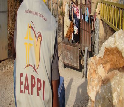 Eine Person mit Hut von hinten, trägt eine Jacke mit der Aufschrift EAPPI und beobachtet das Geschehen.
